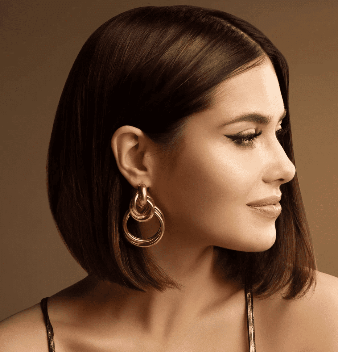 Woman with bob hairstyle and gold hoop earrings gazing sideways on a brown background.