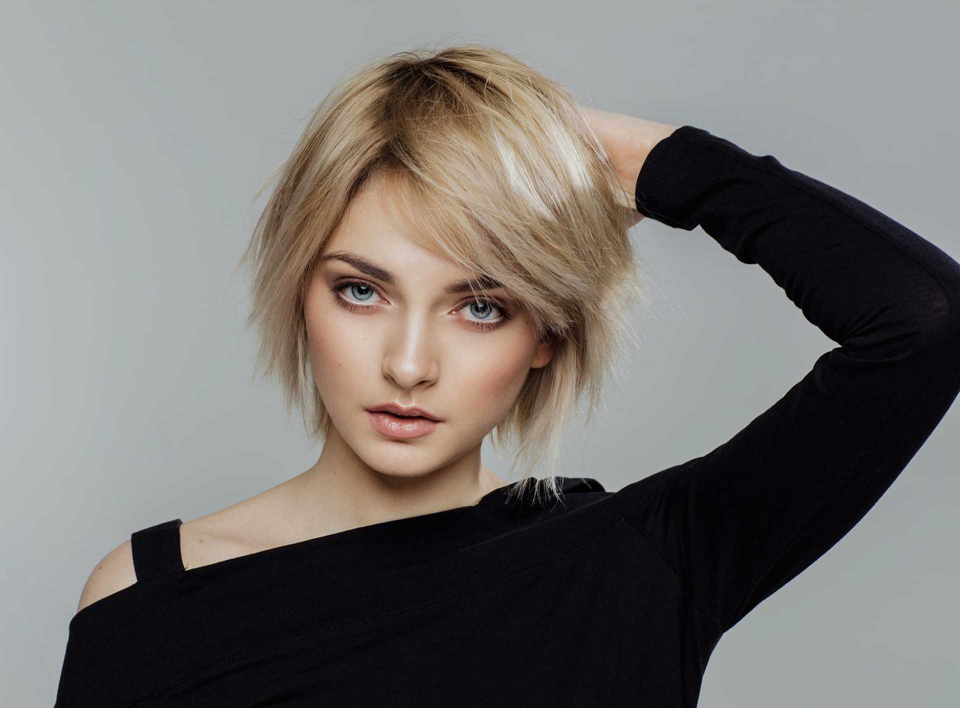 Short-haired woman in black top poses with hand on head against gray background.